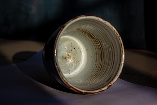 Auf dem Bild ist eine Fotografie der handgemachten Keramiktasse aus dem Onlineshop lebenauslehm.de - Die Tasse trägt den Namen Artium. Die Keramiktasse liegt auf dem Boden, sodass man in die Tasse hineinblicken kann. Dadurch werden die Pinselstriche der Keramikglasur deutlich. Die Tasse steht auf einer weißen Oberfläche.