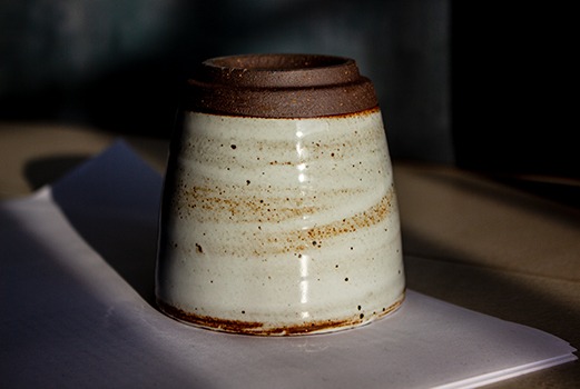 Auf dem Bild ist eine Fotografie der handgemachten Keramiktasse aus dem Onlineshop lebenauslehm.de - Die Tasse trägt den Namen Artium. Die Tasse ist auf den Kopf gestellt und steht auf eine weißen Oberfläche. Hierdurch kommt die untere Seite der Keramiktasse zur Geltung. Hierzu muss man wissen: die Tasse ist Teilglasiert. Das bedeutet, dass etwa 1/6 des unteren Bereiches eine rauhe Oberfläche hat.