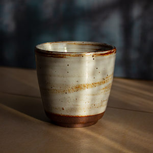 Auf dem Bild ist eine Fotografie der handgemachten Keramiktasse aus dem Onlineshop lebenauslehm.de - Die Tasse trägt den Namen Artium und ist von der Seite fotografiert. Auf die Tasse scheinen die Strahlen der Morgensonne und im Hintergrund erzeugen die Schatten elegante Muster.